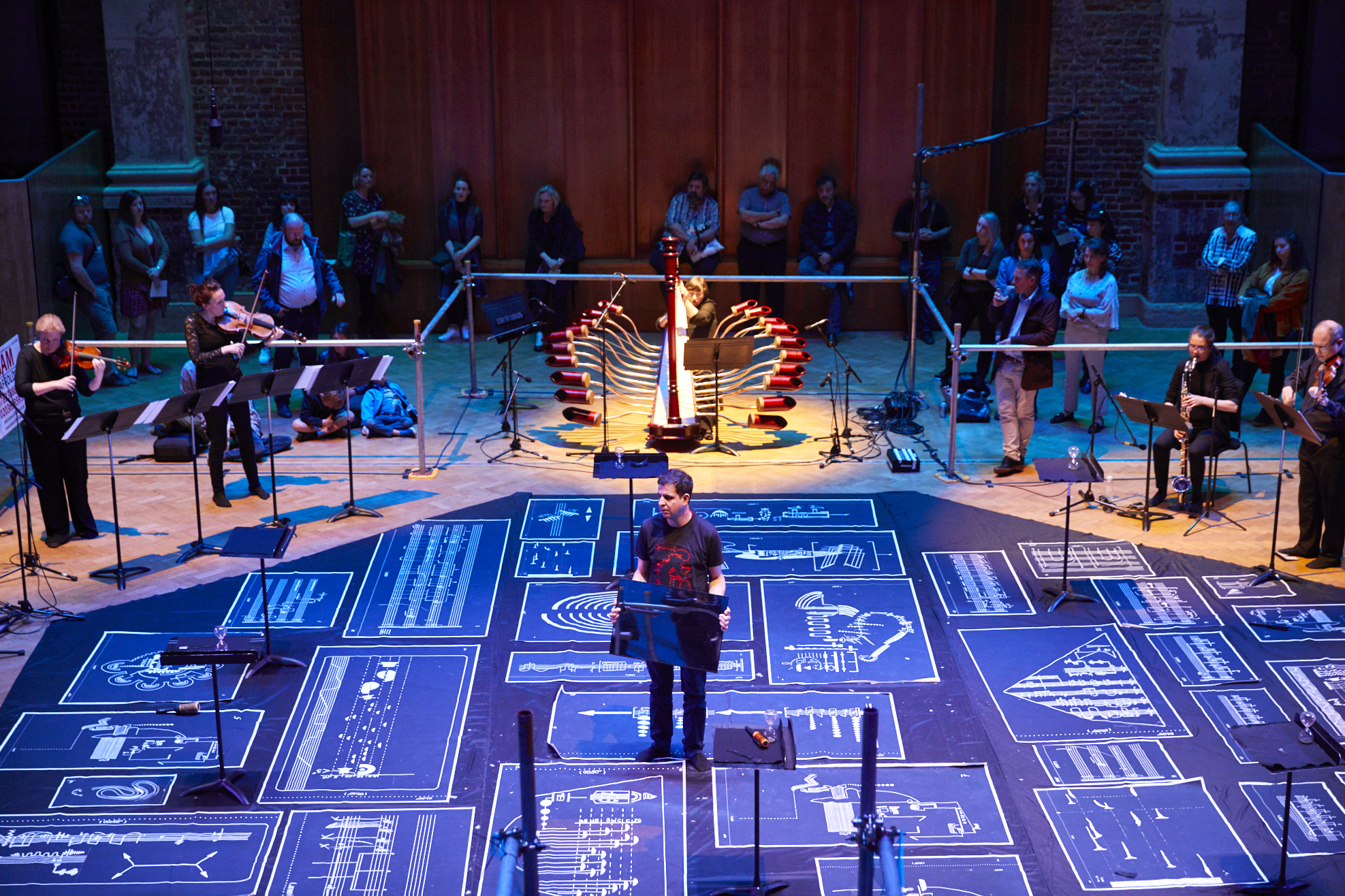 It is a photo of slider showing Davood standing while holding a mirror in the middle of a concert hall